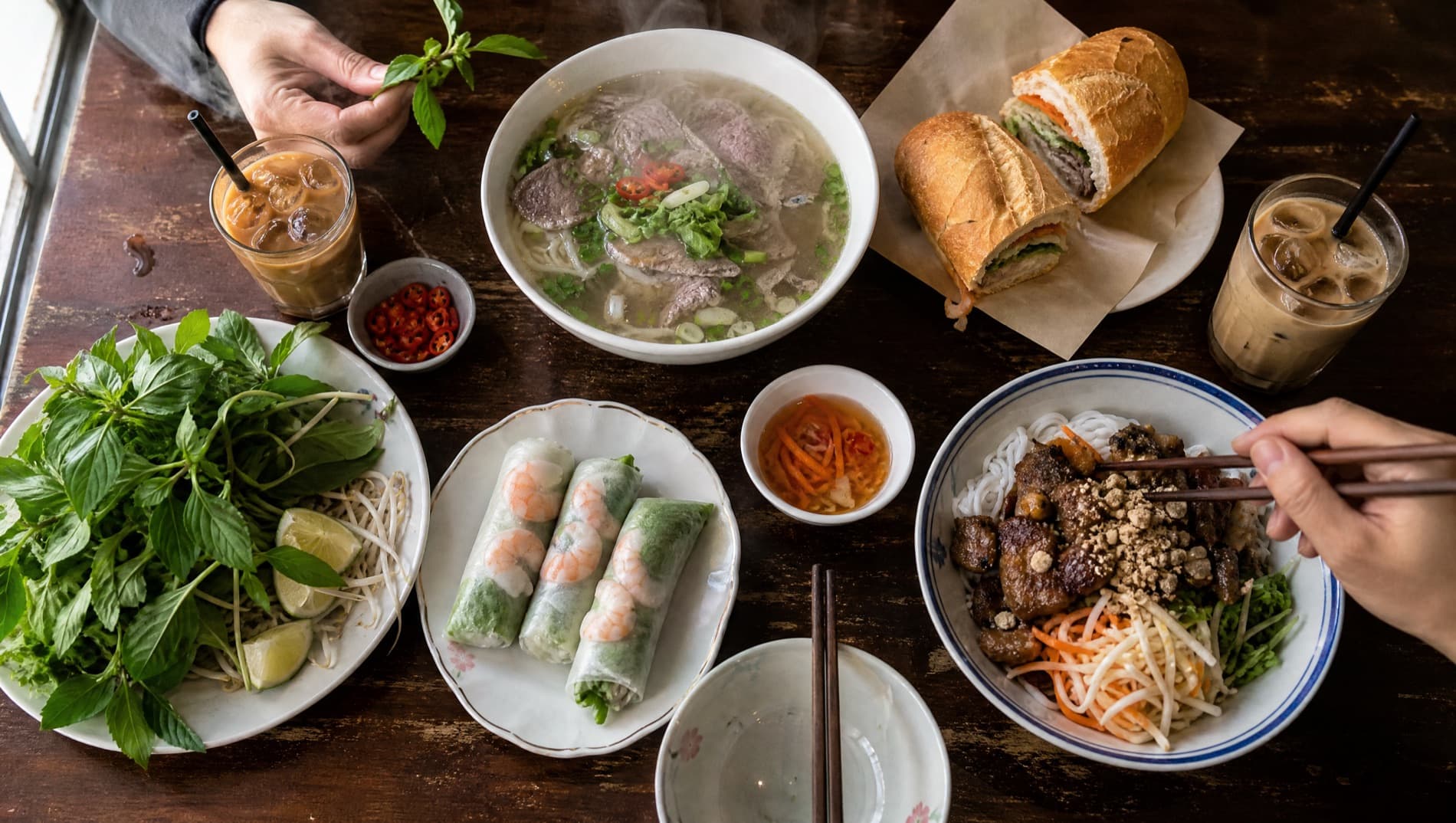 A Vietnamese table set with phở, bánh mì, fresh rolls, a bún bowl, iced coffee and a plate of herbs